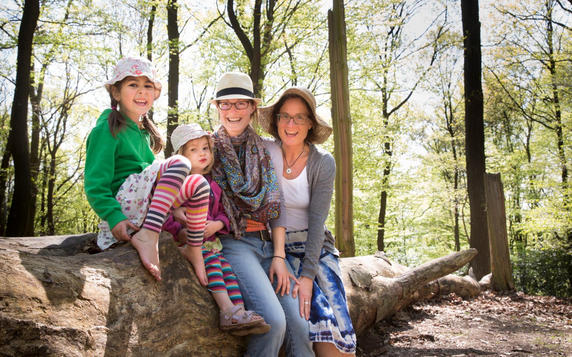 Mütter mit Kindern im Wald. Alle tragen einen Hut und sitzen auf einem Baumstamm.